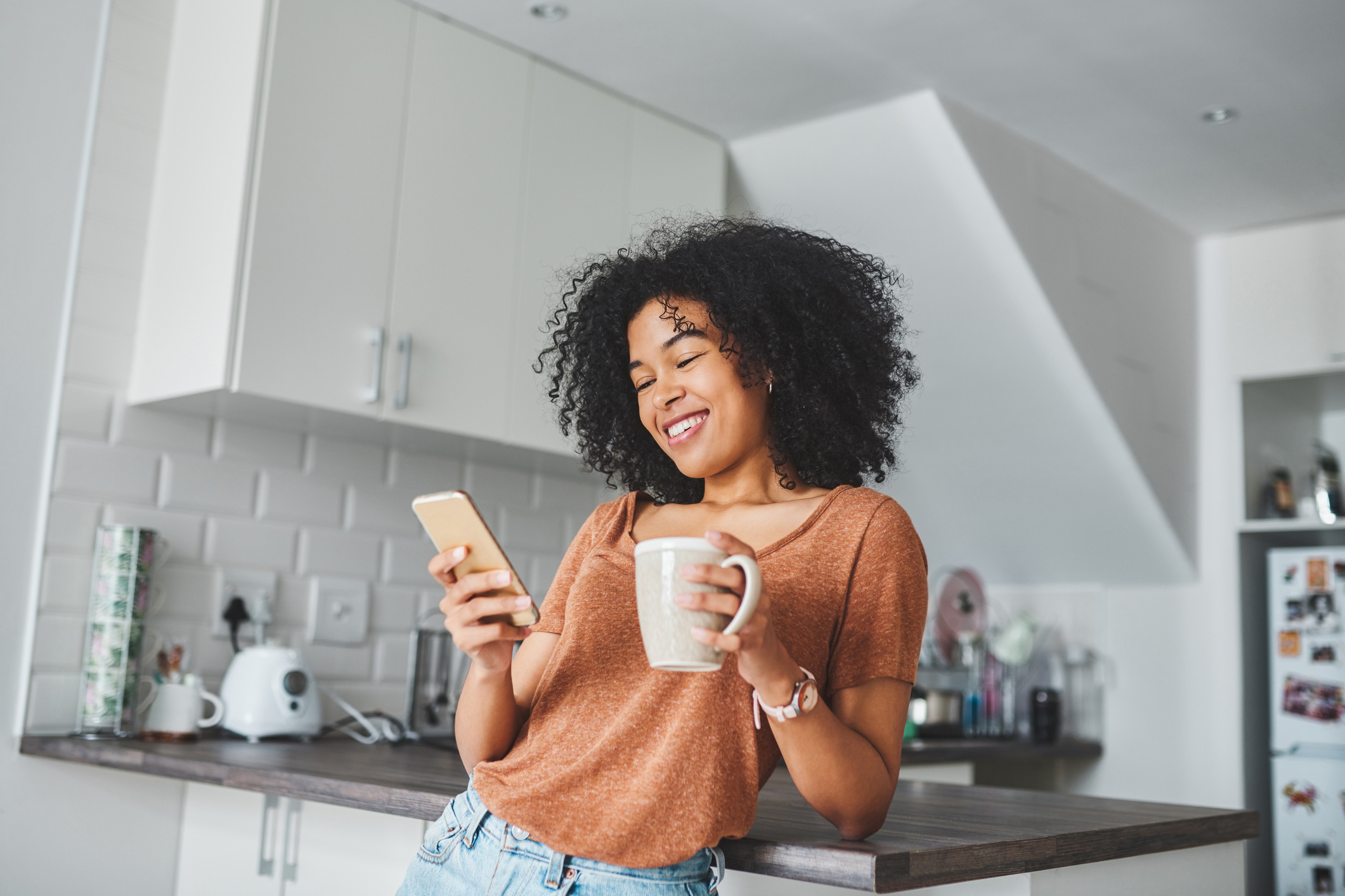 Shot of a young woman using a smartphone to login and search for rental homes, as she is having coffee in the kitchen at home.