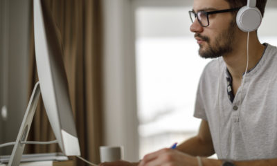 Young man wearing headphones sitting at computer desk taking notes.